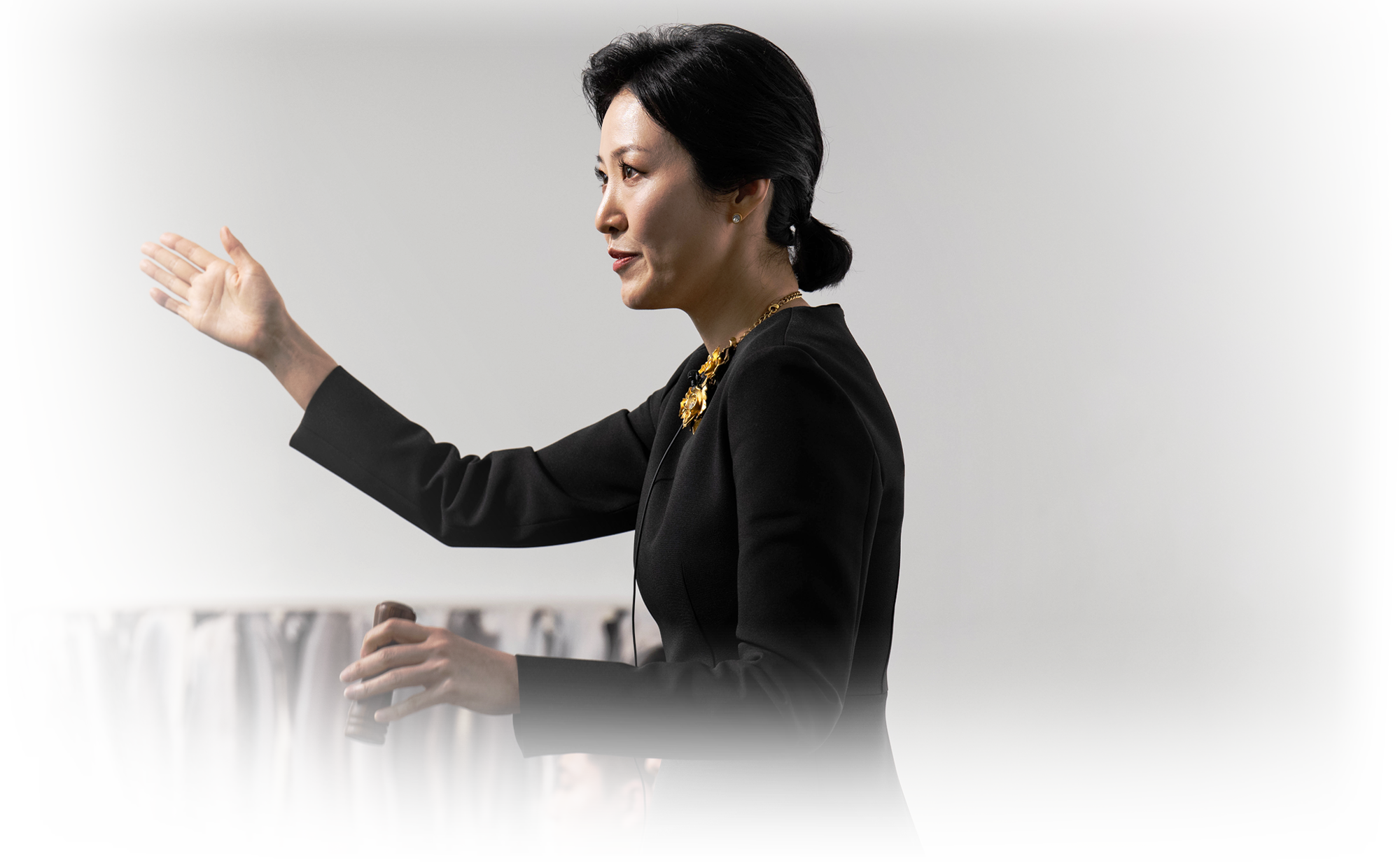 
   			A female auctioneer wearing a black suit is standing and extending her hand toward the audience. 검은 정장을 입은 한 여성 경매사가 서서 관중을 향해 손을 뻗고 있다. 